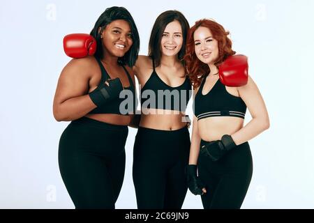 Groupe de trois boxeurs féminins se posant ensemble dans des vêtements de sport et des gants de boxe rouges sur fond blanc. Femelles multiraciales de tailles différentes Banque D'Images