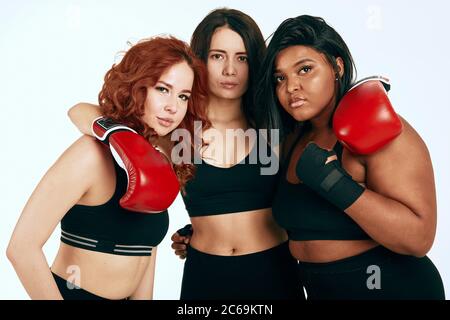 Groupe de filles offensé de différentes tailles et courses, en gants de boxe rouges et vêtements de sport noirs se serrant les uns les autres et regardant sérieusement l'appareil photo Banque D'Images