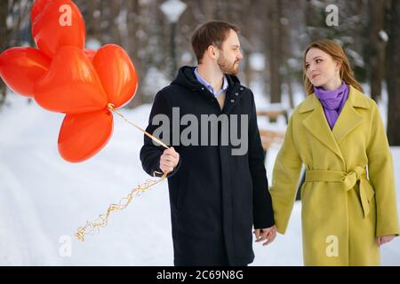 jeune et beau couple caucasien ayant la promenade. romantique homme et femme en manteaux datant, tenir les ballons d'air rouges dans les mains, ils semblent heureux togethe Banque D'Images