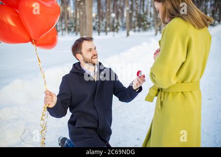 homme barbu en manteau faisant la proposition de mariage à la petite amie dans la rue d'hiver, asseyez-vous sur les genoux et regardez la femme. homme tenir une petite boîte rouge flockée et r Banque D'Images