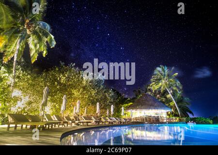 Piscine illuminée de nuit sous une lumière laiteuse, étoiles. Palmiers et parasols fermés avec palmiers sur la piscine à débordement Banque D'Images