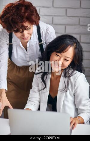 Souriantes femmes asiatiques et caucasiennes travaillant ensemble au bureau avec un ordinateur portable, équipe réussie, partenariat multiethnique Banque D'Images