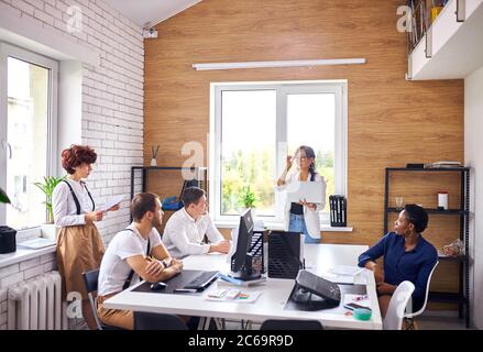 Un jeune leader asiatique couronné de succès portant des vêtements blancs et tenant un ordinateur portable prononce un discours pour un groupe de personnes multiethniques Banque D'Images