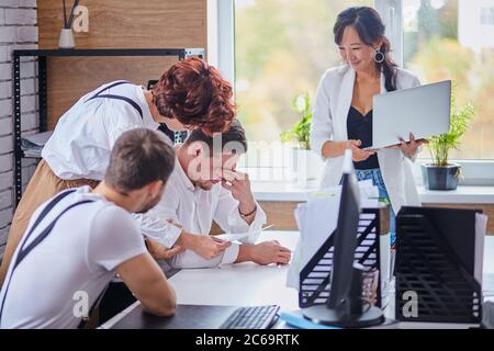 Une équipe de jeunes partenaires d'affaires multiethniques s'assoit Écoutez le succès de l'expression femme asiatique expérimentée qui tient un ordinateur portable blanc. Africain, caucasien et Banque D'Images