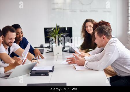 Les jeunes gens qui réussissent en vêtements blancs s'assoient aiment travailler ensemble, rire, sourire. Encouragez les succès au travail, les documents sur table, le bureau intérieur blanc Banque D'Images