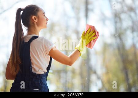 Vue latérale sur une jeune femme du caucase qui lave une fenêtre panoramique, tenant un chiffon rose Banque D'Images