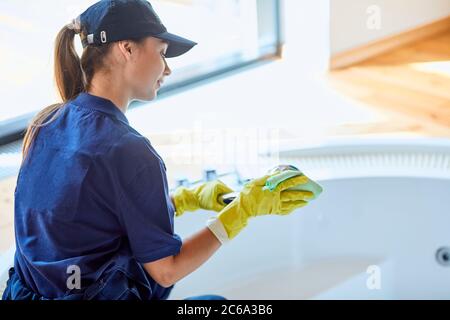 Jeune caucasien concierge en uniforme bleu et gants jaunes bain de nettoyage, poignée de douche Banque D'Images