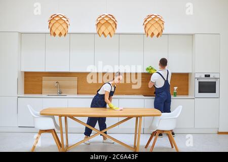 Les nettoyants caucasiens en bleu uniforme essuyant la poussière de la table de cuisine et du placard. Intérieur européen blanc de la cuisine. Concept de service de nettoyage Banque D'Images