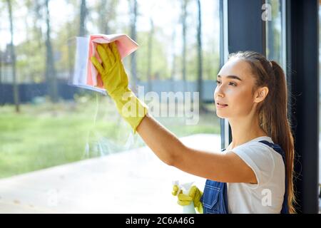 Jeune fille caucasienne en uniforme, sur ses mains gants de protection en caoutchouc jaune. Vaporiser du détergent sur le verre et essuyer la fenêtre avec un chiffon rose. Nettoyage s Banque D'Images