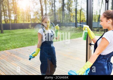 Service de nettoyage fille en uniforme porter des gants de protection jaunes laver la fenêtre à l'extérieur, regarder la réflexion d'elle-même dans le verre Banque D'Images