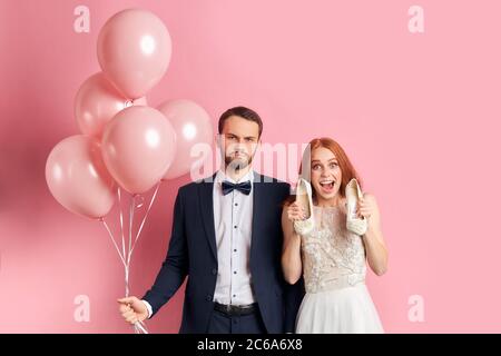 Bonne femme attrayante avec auburn cheveux sourire regardant l'appareil photo et tenant des chaussures dans les mains et sérieux beau homme dans tuxedo. Portrait Banque D'Images