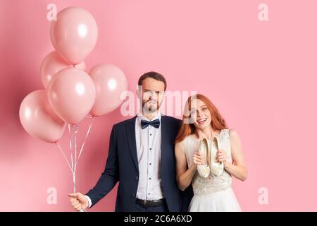 Bonne femme attrayante avec auburn cheveux sourire regardant l'appareil photo et tenant des chaussures dans les mains et sérieux beau homme dans tuxedo. Portrait Banque D'Images