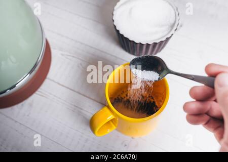 Café du matin - la main d'un homme verse du sucre dans un café moulu dans une tasse jaune Banque D'Images