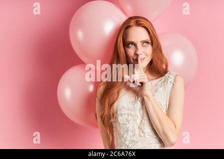 Belle femme avec les cheveux auburn portant robe blanche avec ballons d'air derrière, penser, regarder. Isolé sur fond rose Banque D'Images