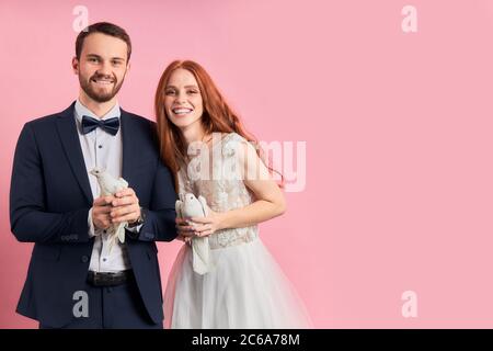 Belle femme avec les cheveux rouges et homme portant tuxedo tenant des pigeons dans les mains, heureux ensemble, stand isolé sur fond rose . Symbole colombe de h Banque D'Images