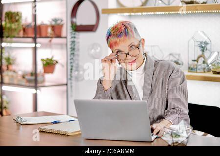 Une femme âgée confiante portant des lunettes est assise sur un ordinateur portable dans une salle de bureau blanche, des carnets et des papiers sur une table. Femme mature vêtue de blazer, avec Banque D'Images