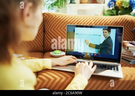 Vue de derrière une femme étudier en ligne sur un ordinateur portable avec un enseignant utilisant la vidéo appel dans le salon en journée ensoleillée. Banque D'Images