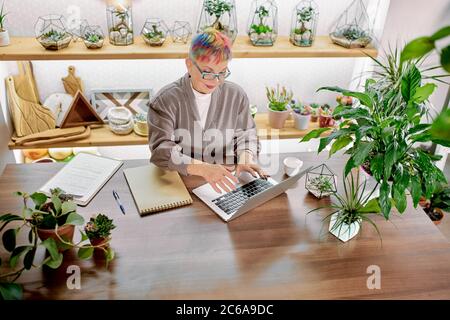 Femme caucasienne senior en lunettes, avec une coiffure moderne colorée, profitez de travailler au bureau, sourire au travail. Utilisation d'un ordinateur portable Banque D'Images