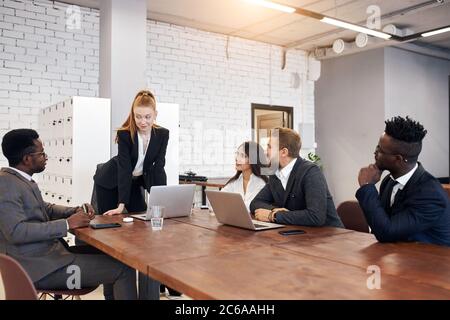 Jeune femme caucasienne aux cheveux rouges vêtue d'un costume formel tenant une réunion avec un groupe multiethnique de personnes impliquées dans le commerce. Concept d'entreprise Banque D'Images