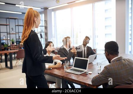 Jeune femme caucasienne aux cheveux rouges vêtue d'un costume formel tenant une réunion avec un groupe multiethnique de personnes impliquées dans le commerce. Concept d'entreprise Banque D'Images