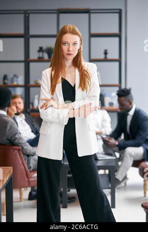 Belle femme sérieuse dans blazer blanc et avec les cheveux rouges, regarder la caméra pendant la réunion d'affaires, processus de négociations de peopl africain et caucasien Banque D'Images