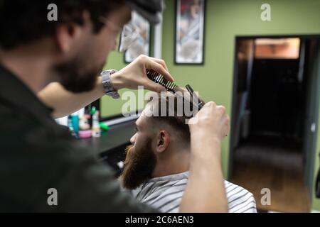 Jeune homme barbu se coupe par coiffure while sitting in chair at barbershop Banque D'Images