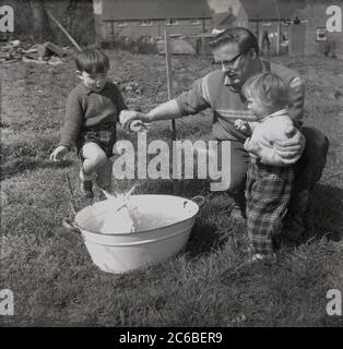 années 1950, historique, à l'extérieur dans une zone d'un jardin avec wirefencing, un père avec ses deux jeunes enfants.. Qui sont excités de voir leur lapin animal de compagnie ayant un lavage dans une petite baignoire à linge en métal, Angleterre, Royaume-Uni. Banque D'Images
