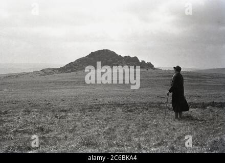 Années 1940, historique, une marcheuse mature, avec bâton, debout sur un sol plat regardant le paysage à Higher White Tor, Dartmoor, Devon, Angleterre, Royaume-Uni. L'un des tors supérieurs autour de la zone des deux ponts de la lande, situé sur une crête d'une colline signifie une vue magnifique. Les tors sont des affleurements de granit et le parc national de dartmoor en compte plus de 160. Banque D'Images