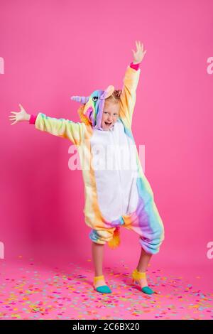 Bonne petite fille dans kigurumi unicorn sur fond rose se réjouit dans les confettis multicolores, espace pour le texte Banque D'Images