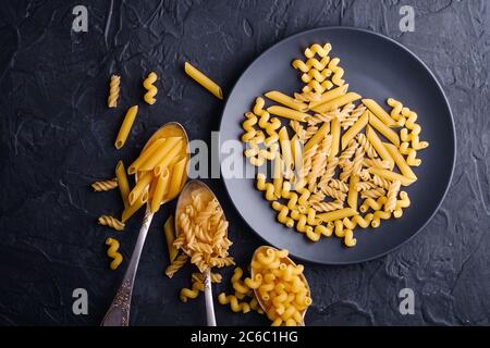 Trois cuillères à couverts et assiette avec variété de pâtes de blé doré non cuites sur fond noir foncé texturé, vue du dessus Banque D'Images