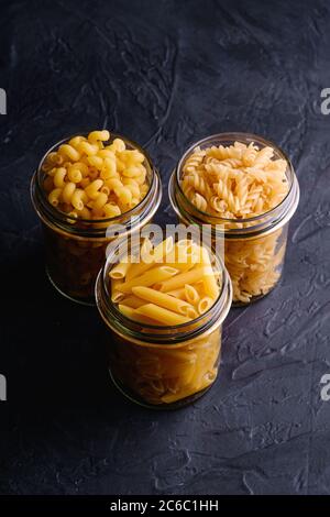 Trois pots en verre avec variété de pâtes de blé doré non cuites sur fond noir foncé texturé, vue en angle Banque D'Images