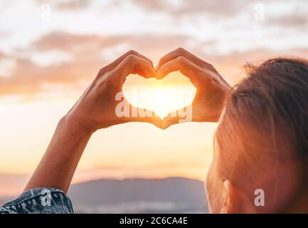 Jeune femme en forme DE COEUR avec ses doigts attrapant les poutres de coucher de soleil avec un fond spectaculaire de ciel. Image de concept d'amour ou d'homme et de nature. Banque D'Images
