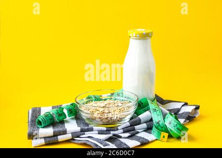 Flocons d'avoine dans un bol, lait dans une bouteille en verre et ruban vert centimètre sur fond jaune vif avec serviette de cuisine, espace de copie Banque D'Images