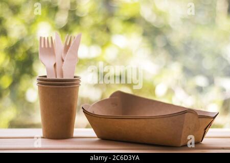 Fourchettes en bois et couteaux dans des tasses artisanales, assiettes en papier, fond vert naturel flou. Ustensiles de cuisine jetables écologiques. Écologie, zéro déchet Banque D'Images