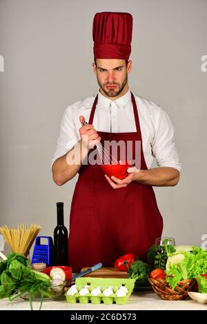 Chef avec un visage sérieux tient un bol et fouetter sur fond gris. Cuisine et concept de cuisine. Homme dans le chapeau de cuisinier et le tablier tient les ustensiles de cuisine. Cuisine travaille dans la cuisine près de la table avec des légumes et de la nourriture Banque D'Images