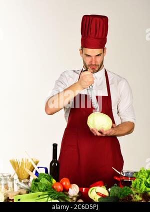 Chef avec le visage strict met le couteau dans le chou sur fond blanc. L'homme en chapeau et en tablier contient des ingrédients. Cuisiner travaille dans la cuisine près de la table avec des légumes et des outils. Ustensiles de cuisine et concept de cuisine. Banque D'Images