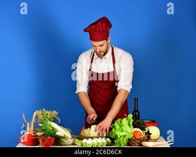 Homme dans le chapeau de cuisinier et le tablier coupe le chou. Chef avec chops de visage très occupés chou avec couteau sur fond bleu. Cuisiner travaille dans la cuisine près de la table avec des légumes et des outils. Ustensiles de cuisine et concept de cuisine. Banque D'Images