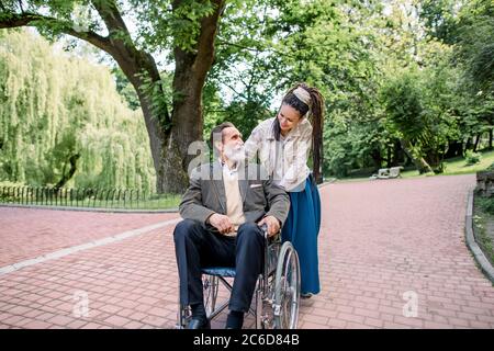 Homme barbu senior handicapé en fauteuil roulant et son assistant de fille créative avec des dreadlocks, en vêtements de taille basse, marchant dans le parc par beau temps. Fille Banque D'Images