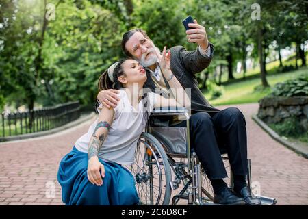 Fille de Hipster avec de longues anses et beau barbu homme principal grand-père en fauteuil roulant au parc prenant selfie. Mode de vie des personnes handicapées. Soins Banque D'Images