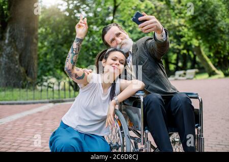 Portrait d'un beau barbu senior sur fauteuil roulant et de son jeune boxeur ou petite-fille avec tatouage et dreadlocks, faisant selfie et Banque D'Images