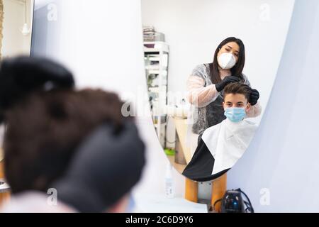 Miroir montrant un coiffeur avec un masque de protection peignant un garçon avec un masque assis sur une chaise dans un salon de coiffure Banque D'Images