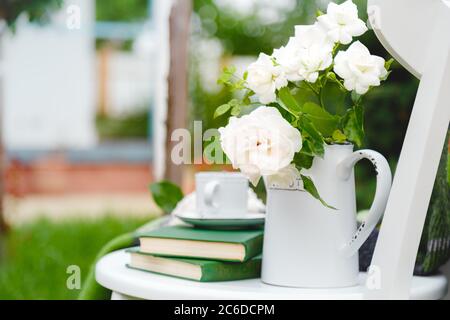 Fleurs rose sauvage blanche dans vase théière, tasse de thé, livres sur chaise en bois blanc dehors dans le jardin ensoleillé. Romantique provence petit déjeuner de loisirs sur confortable Banque D'Images