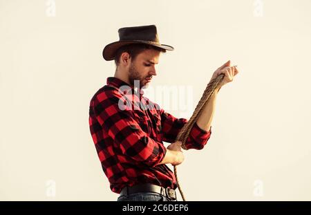 Ouvrier de Ranch. Éco-exploitation. Concept agricole. Bel homme avec chapeau et tenue de style rustique. Garder le ranch. Un agriculteur réfléchi qui réfléchit aux affaires. La vie au ranch. Cowboy avec lasso corde ciel fond. Banque D'Images