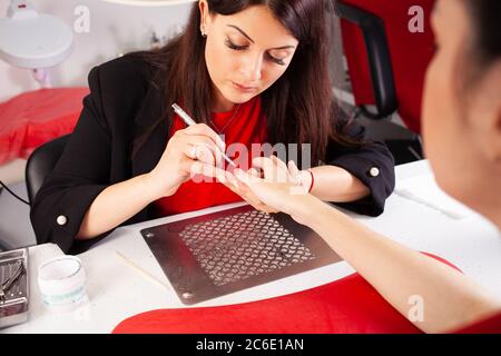 La femme lubrifie les ongles du client avec une brosse. Le processus de manucure dans un salon de beauté. Salon en noir, rouge, blanc. Le maître de manucure pu Banque D'Images