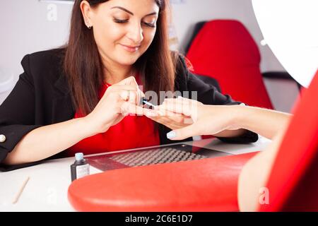 La femme couvre les ongles du client avec du vernis bleu. Le processus de manucure dans un salon de beauté. Salon en noir, rouge, blanc. Le maître de manucure Banque D'Images