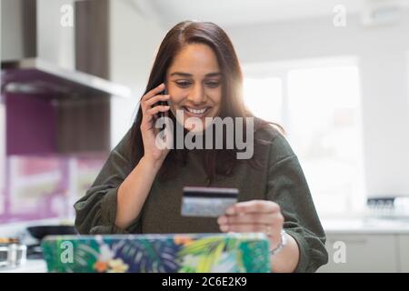 Femme souriante avec carte de crédit et smartphone payant des factures dans la cuisine Banque D'Images