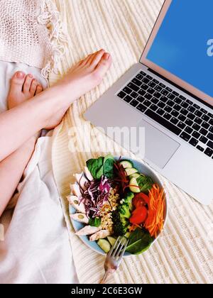 Par dessus une femelle anonyme assise pieds nus sur un lit souple avec un netbook et une salade saine tout en étant à la maison Banque D'Images