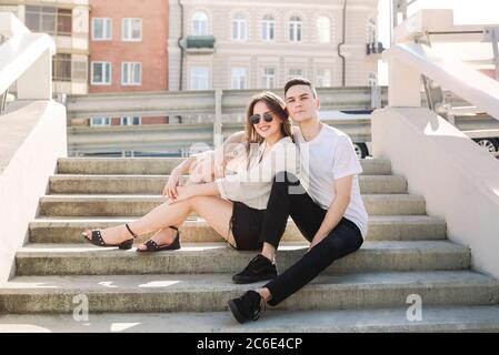 Petite amie et petit ami élégants qui s'embrasse et s'assoit dans les escaliers en pierre dans la rue par beau temps, tout en regardant l'appareil photo Banque D'Images