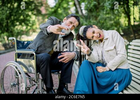 Portrait de jeune fille moderne avec des dreadlocks, assis sur un banc, avec l'homme handicapé en fauteuil roulant, posant à la caméra dans le parc Banque D'Images