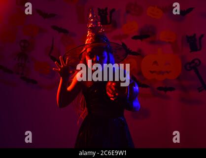 Un enfant en costume de sorcières effrayantes tient une citrouille ou une lanterne. Fille avec visage sinistre sur fond rouge sanglant avec chauves-souris et décor de citrouilles. Petite sorcière portant un chapeau noir. Halloween Party and decorations concept Banque D'Images